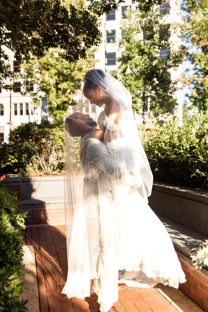 The groom lifts the bride into his arms as her veil cascades dramatically over them both, soft sunlight spilling through the trees. The red brick pathways and urban greenery around Miller Plaza frame this romantic, fairytale-like image.