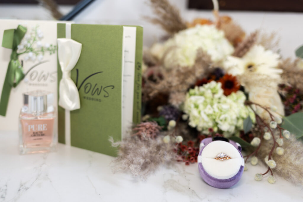 A still-life detail from this Chattanooga farm wedding features two vow books tied with satin ribbons, a bottle of perfume, and a velvet ring box nestled beside a textured bouquet of hydrangeas, pampas grass, and dried florals. The composition reflects both sentimentality and style, hinting at the couple’s earthy, romantic aesthetic.