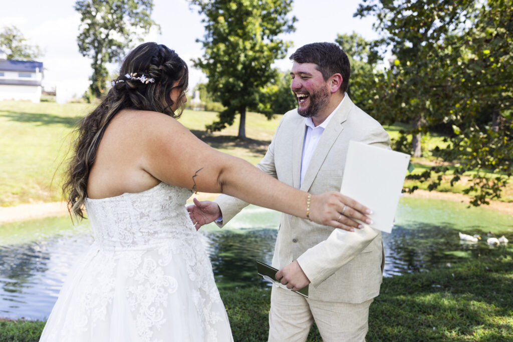 The bride and groom laugh with pure happiness as they move toward each other for a hug beside the pond. He holds a small book for his vows, while she reaches out with open arms, her bouquet still waiting nearby. The scene feels lighthearted and full of connection, a tender reflection of their Candies Creek Farm Wedding day.