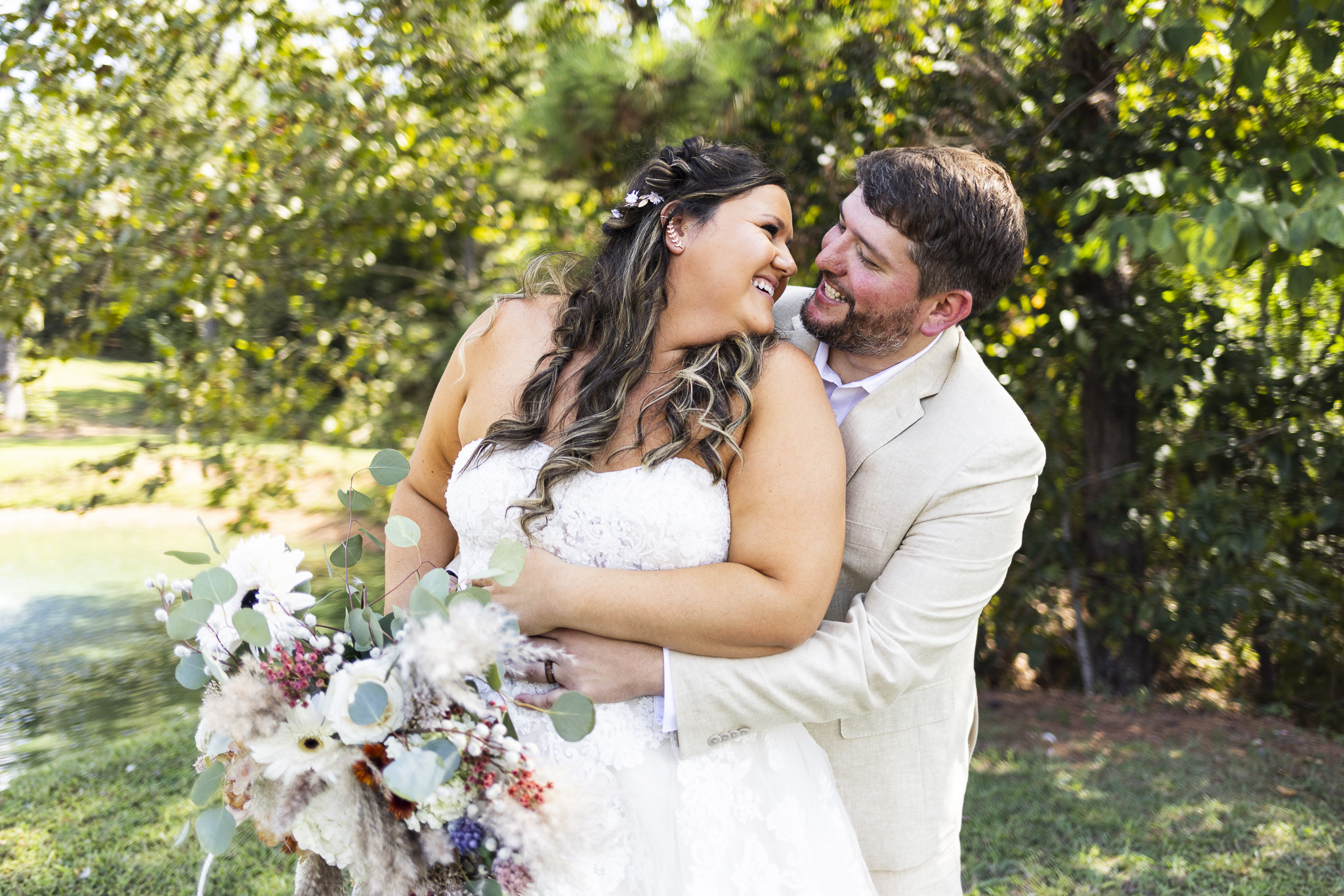 The bride leans back into the groom’s arms, both of them grinning as they share a quiet moment surrounded by trees and sunlight. Her bouquet, filled with pampas grass, muted roses, and eucalyptus, adds soft texture against her lace gown. The natural setting and their laughter make this portrait a quintessential reflection of their Candies Creek Farm Wedding joy.