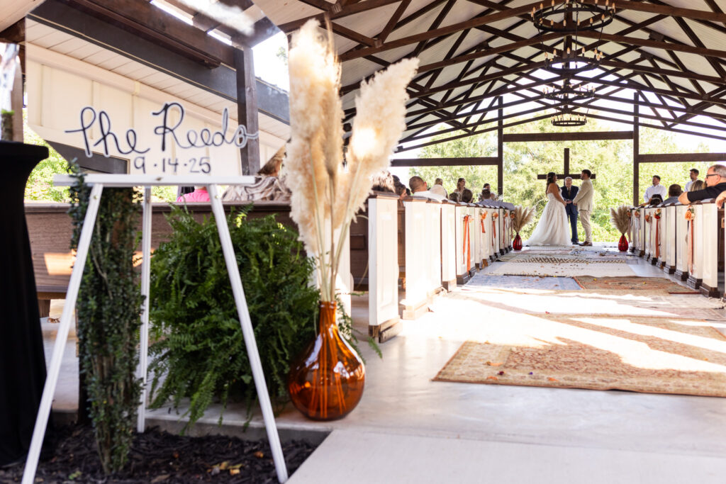 The open-air chapel at this Candies Creek Farm Wedding frames a beautiful view of the ceremony, seen from the aisle’s entrance. A mirrored welcome sign reading “The Reeds 9.14.25” leans against lush ferns, while a line of vintage rugs and pampas arrangements lead toward the couple standing at the altar beneath a wooden cross. The natural light and warm tones set a serene, heartfelt atmosphere.