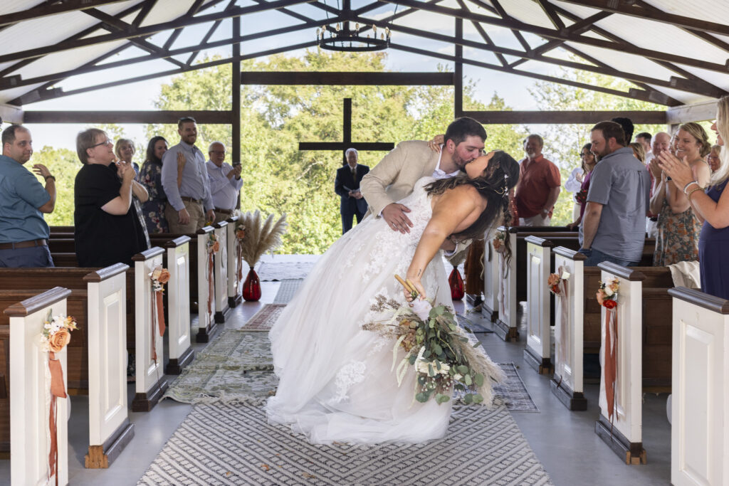 Sunlight pours through the open beams during this Candies Creek Farm Wedding as the newlyweds share a kiss while coming back down the aisle. Guests stand and cheer, some capturing the moment on their phones, while the bride’s pampas-filled bouquet brushes against her lace gown. The groom’s light tan suit complements the warm neutral palette surrounding them, embodying a joyful recessional.