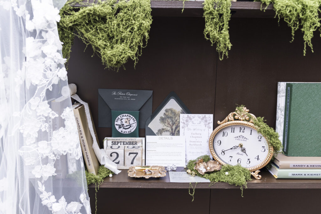 A styled flat-lay vignette from a Lancaster wedding featuring the couple’s stationery suite, a black envelope, a gold-rimmed clock, wedding rings displayed in a vintage tray, and a September 27 date block. Green moss weaves through the setup, uniting the elegant woodland aesthetic of the day.