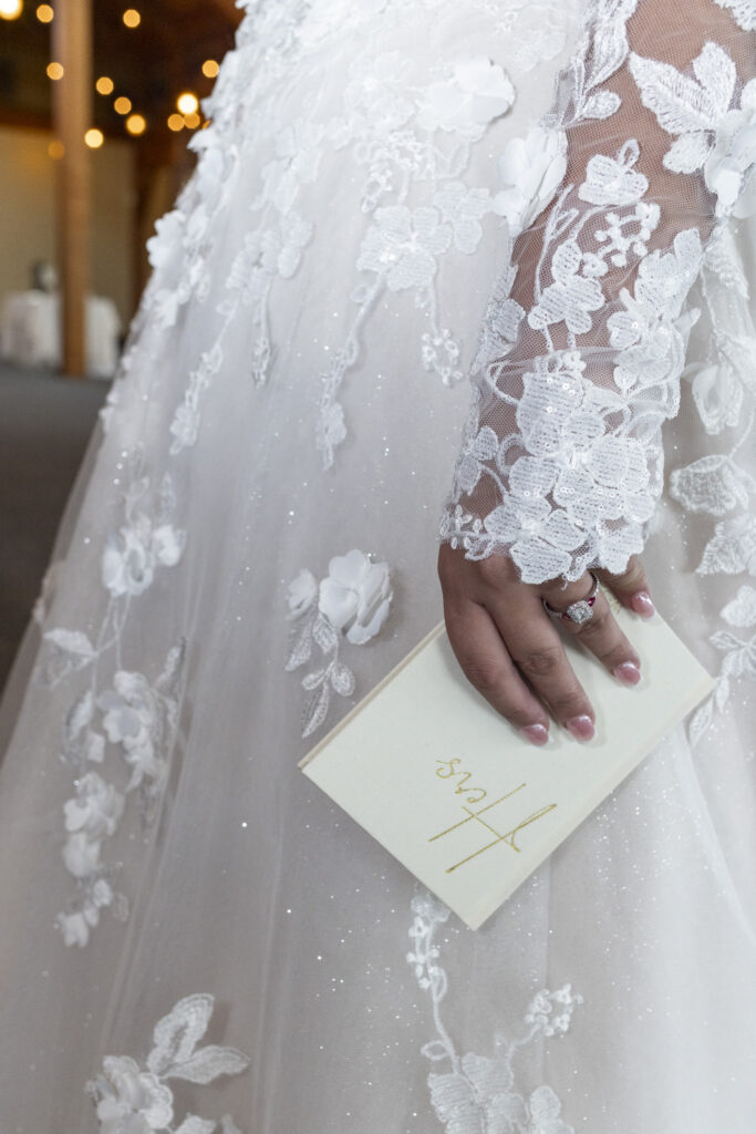 The bride holds her ivory vow book embossed with gold script reading “Hers.” Her hand, adorned with an elegant engagement ring, rests against the detailed floral lace of her gown—an intimate detail capturing bridal elegance and anticipation at this Lancaster wedding.