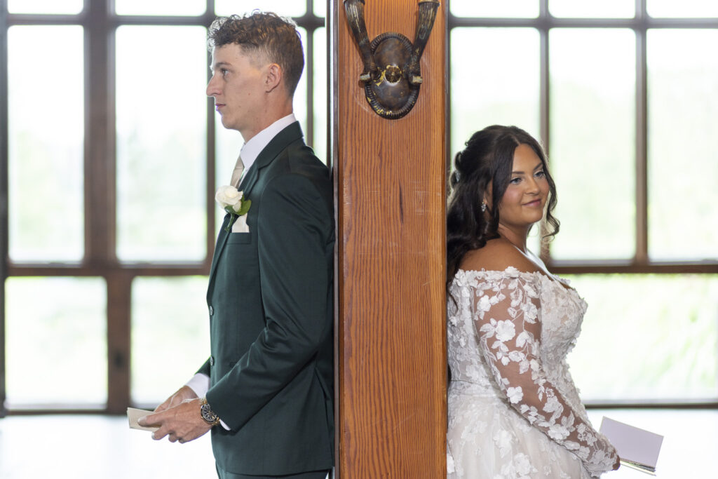 The bride and groom stand back-to-back on either side of a wooden beam, each holding their vow books, sharing a private pre-ceremony moment without seeing one another. Soft light from tall windows illuminates them, creating an emotional and intimate pause in their Lancaster wedding day.