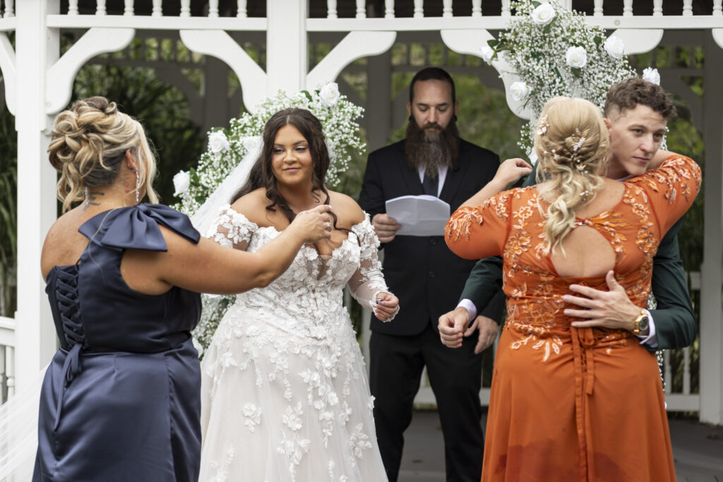 A touching scene at The Lodge at Liberty Forge wedding shows both sets of parents joining the couple at the altar for a blessing. The bride’s mother, in navy satin, adjusts her daughter’s gown while the groom embraces his mother, who wears a burnt orange lace dress.