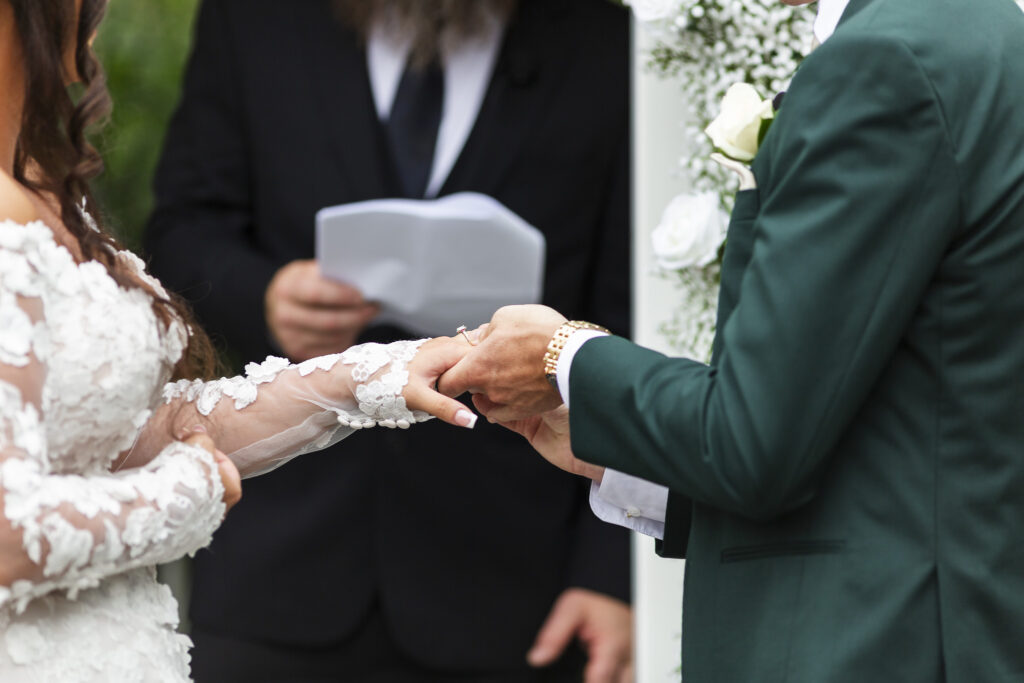 A close-up captures the groom gently placing the wedding ring on his bride’s finger, his gold watch glinting as the officiant stands behind them reading vows. The intricate floral lace of her sleeve and the tender moment emphasize the intimacy of their ceremony at The Lodge at Liberty Forge.
