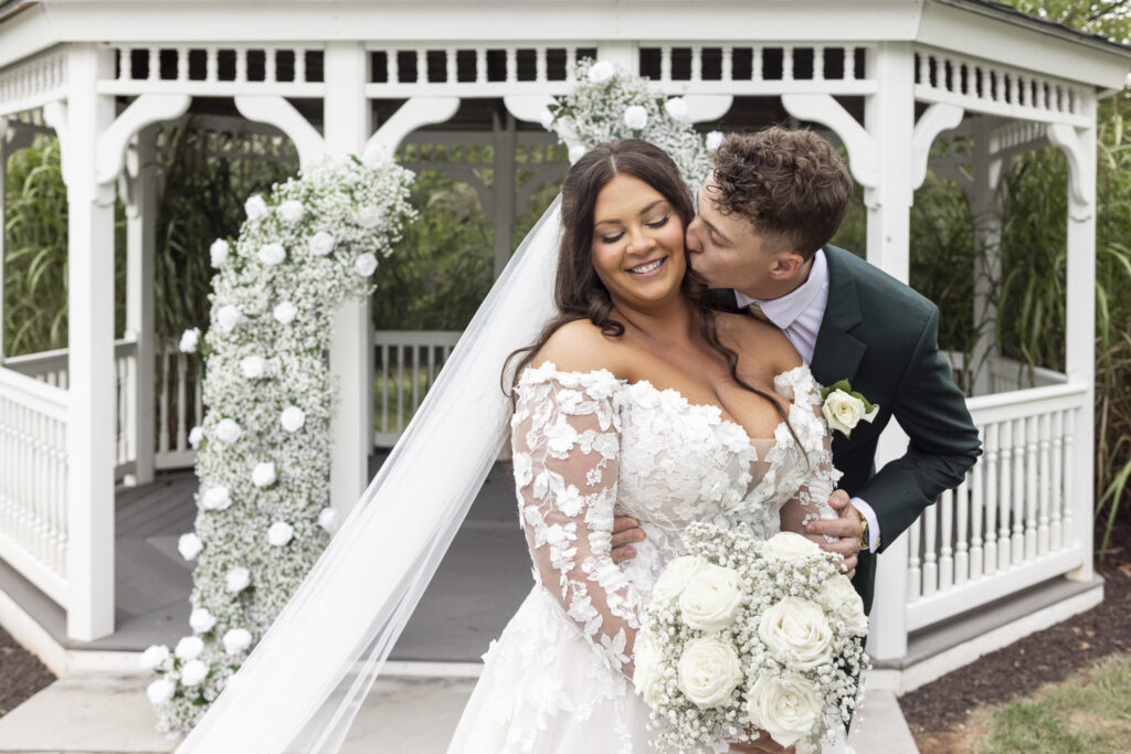 The groom leans in to kiss the bride on the cheek as she smiles softly, standing together in front of the gazebo adorned with white florals. Her bouquet of roses and baby’s breath complements the gentle romance of their wedding at The Lodge at Liberty Forge.
