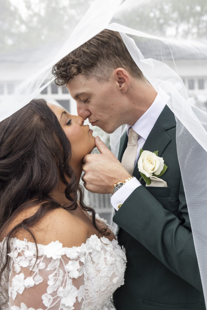 Beneath the bride’s flowing veil, the couple shares an intimate kiss, their foreheads touching tenderly. The soft light filters through the sheer fabric, creating a dreamy, timeless moment from their wedding at The Lodge at Liberty Forge.