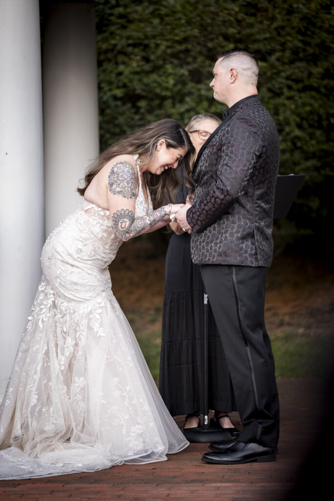 The bride bends forward laughing as she holds her groom’s hands, their playful moment captured beneath the classic white columns of their outdoor Philadelphia wedding ceremony.