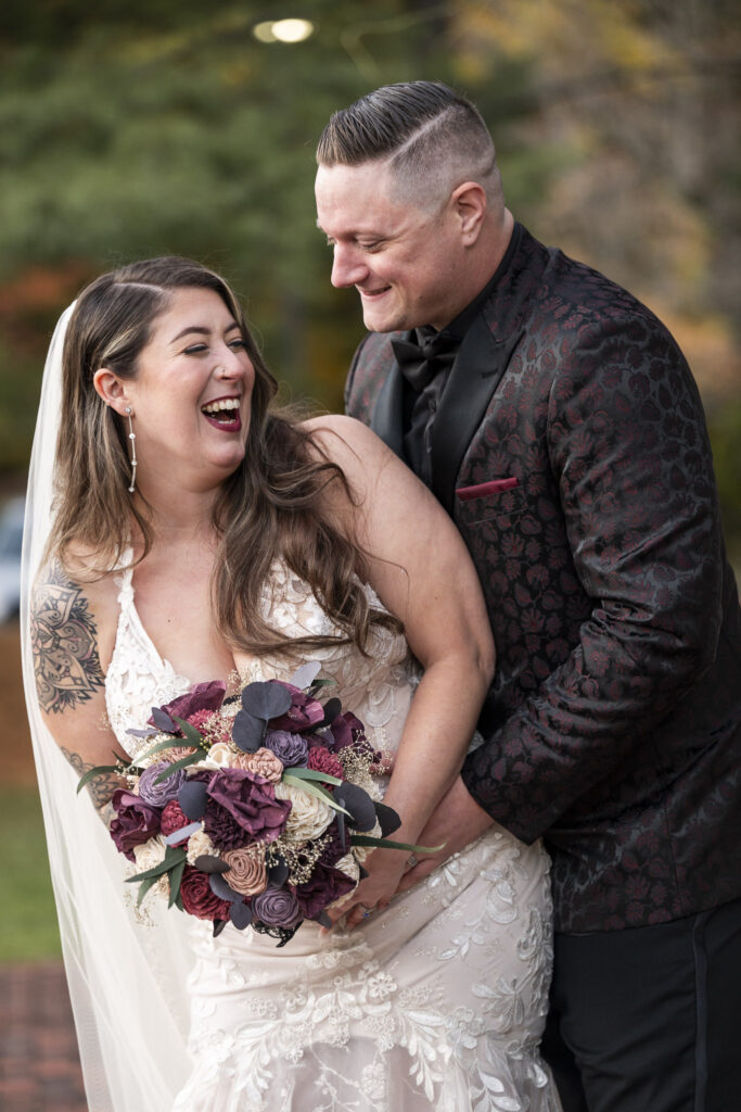 The newlyweds share a candid laugh, their faces full of joy, as the groom wraps his arms around the bride. Her bouquet of plum, ivory, and mauve flowers adds a romantic pop of color to their Philadelphia wedding portraits.