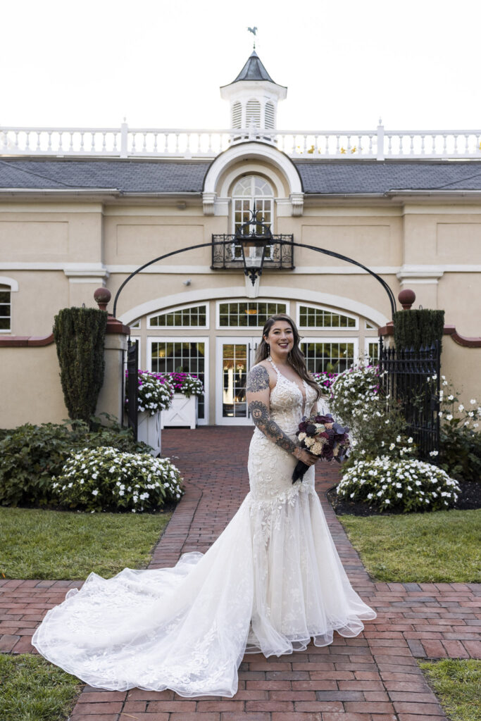 The bride stands in front of the stately Pyn Ryn mansion, her lace gown flowing into a long train over the brick walkway. Her bouquet of rich plum and cream tones complements the lush floral landscaping surrounding the venue’s entrance.