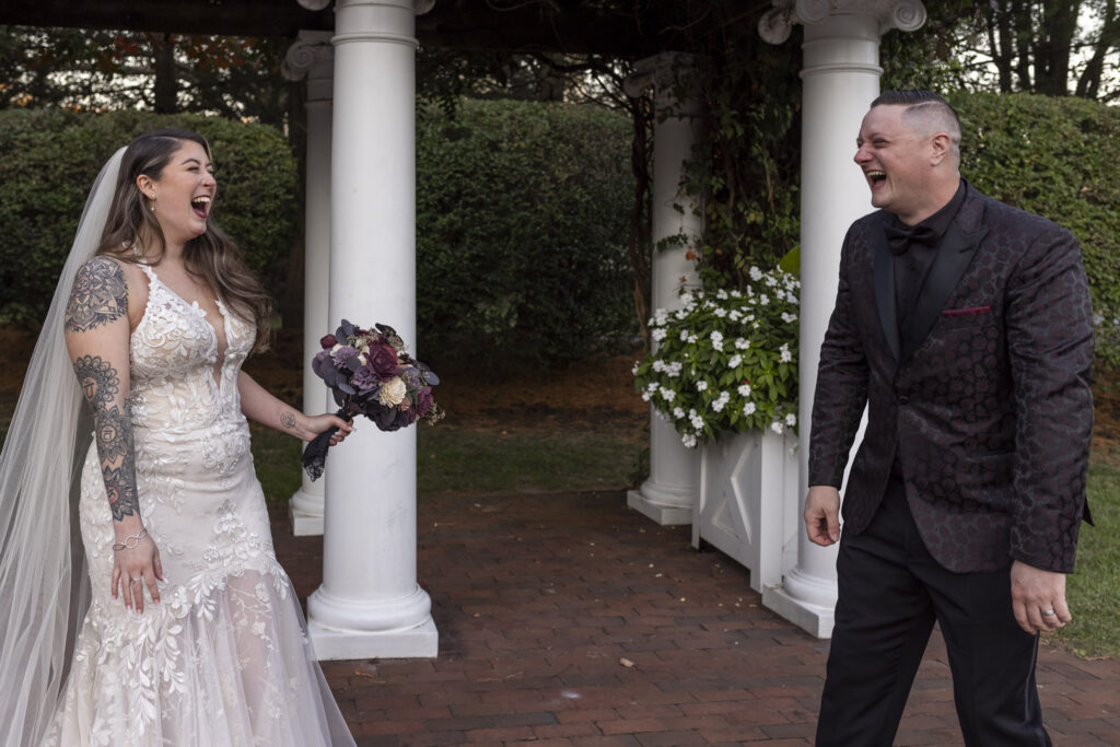 The bride and groom laugh together under the white-columned arbor surrounded by blooming greenery. Their relaxed energy and shared laughter reflect the lighthearted romance of their outdoor Pyn Ryn Wedding celebration.