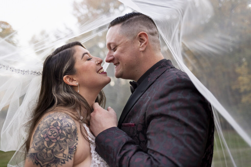 The couple shares an intimate moment beneath the bride’s veil, smiling as their foreheads nearly touch. The groom’s deep burgundy patterned jacket and the soft fall backdrop highlight the romantic atmosphere of their masquerade wedding portraits.