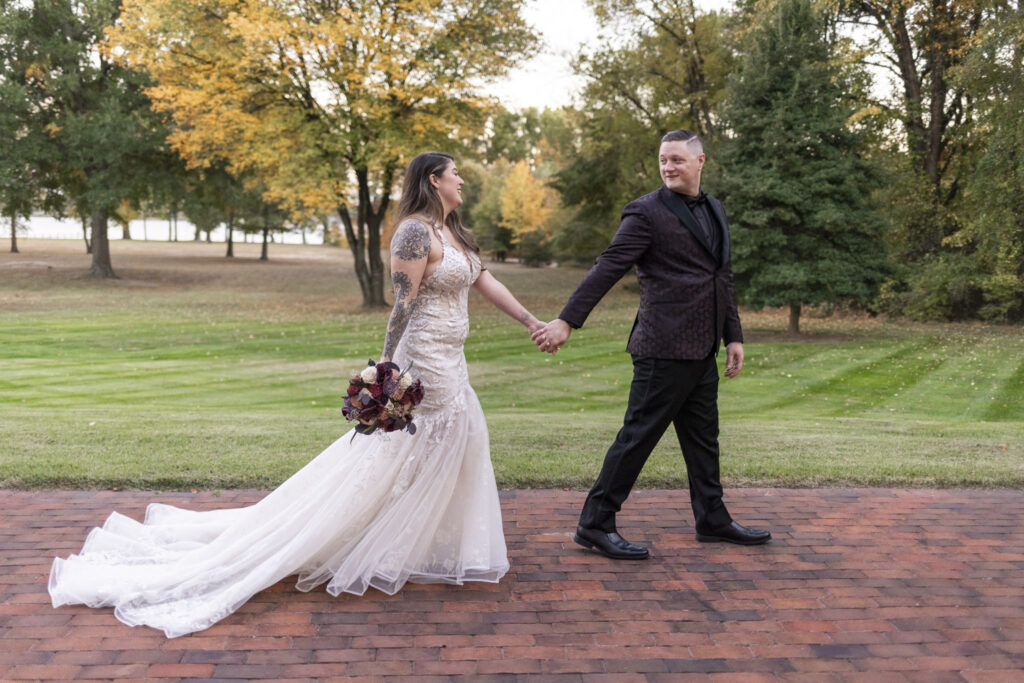 Hand in hand, the couple walks down a red brick path surrounded by autumn trees during their Pyn Ryn wedding day portraits. The bride’s long train trails behind her, and her bouquet of plum and ivory blooms stands out against the golden foliage.