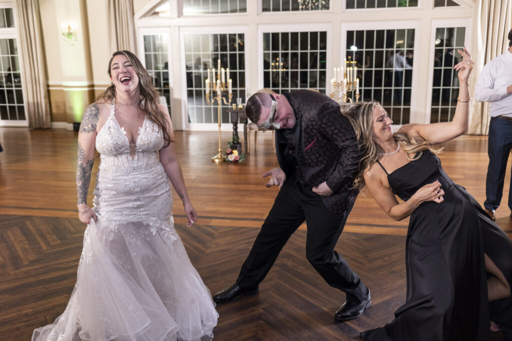 The bride, groom, and a bridesmaid share a carefree dance together, laughing and striking playful poses beneath the ballroom chandeliers. The scene radiates the uninhibited joy of their masquerade wedding reception.