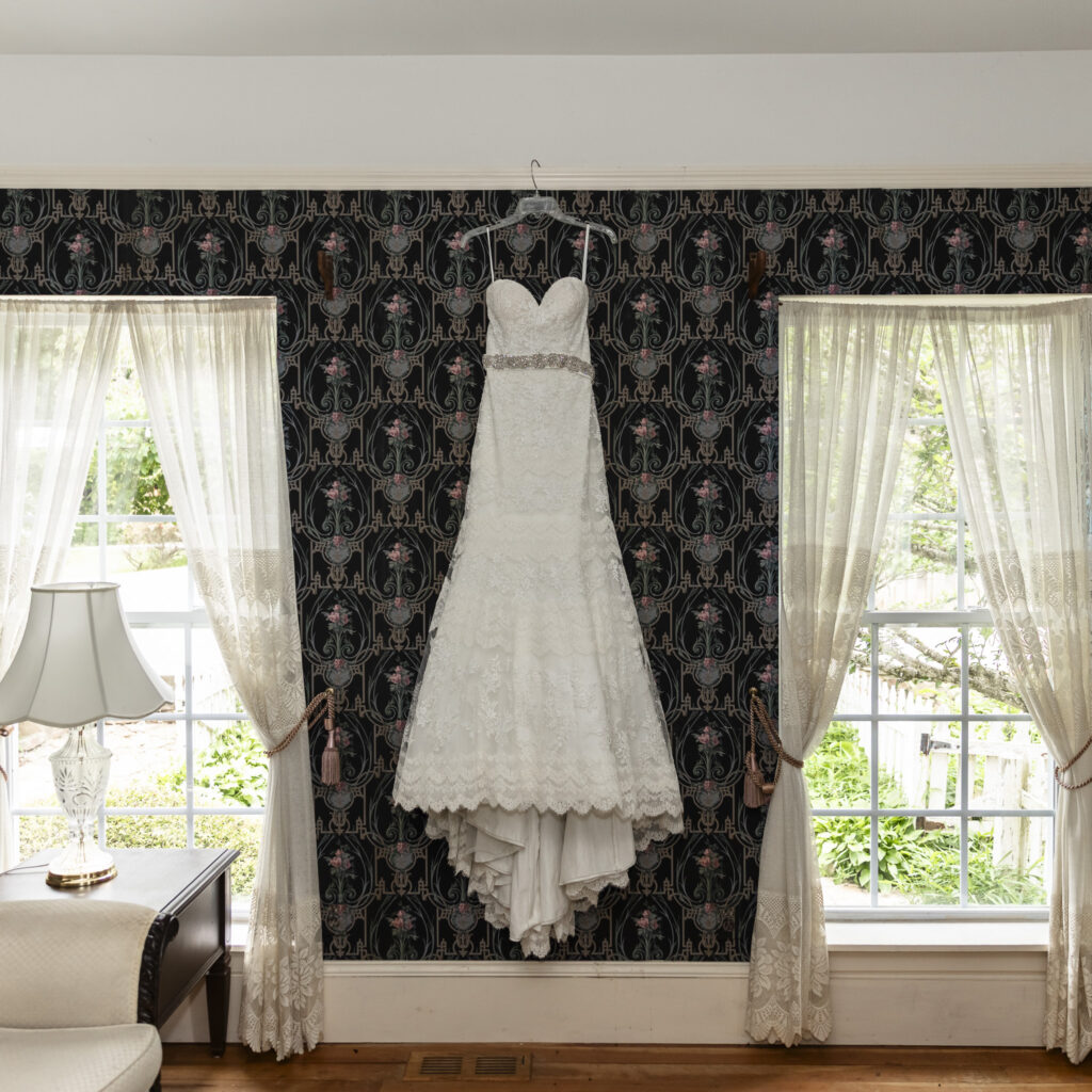 Classic lace wedding dress with sweetheart neckline and jeweled belt hanging between two lace-curtained windows in a vintage room.