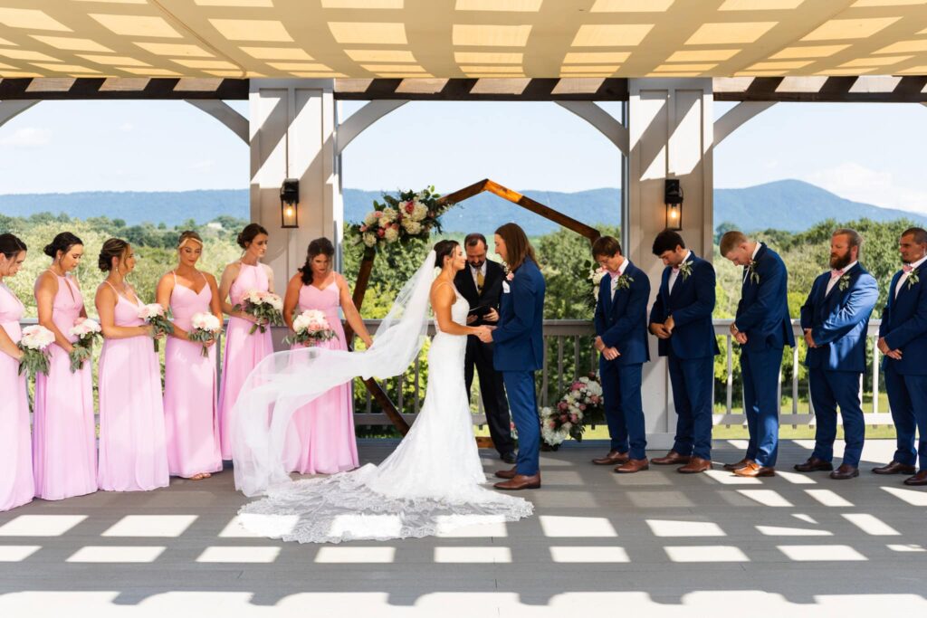 A bright summer ceremony takes place under a shaded pergola overlooking rolling blue mountains, with bridesmaids in pink dresses and groomsmen in blue suits surrounding the couple as the veil lifts in the breeze, showing what time do weddings usually start for warm-weather weddings with full daylight.
