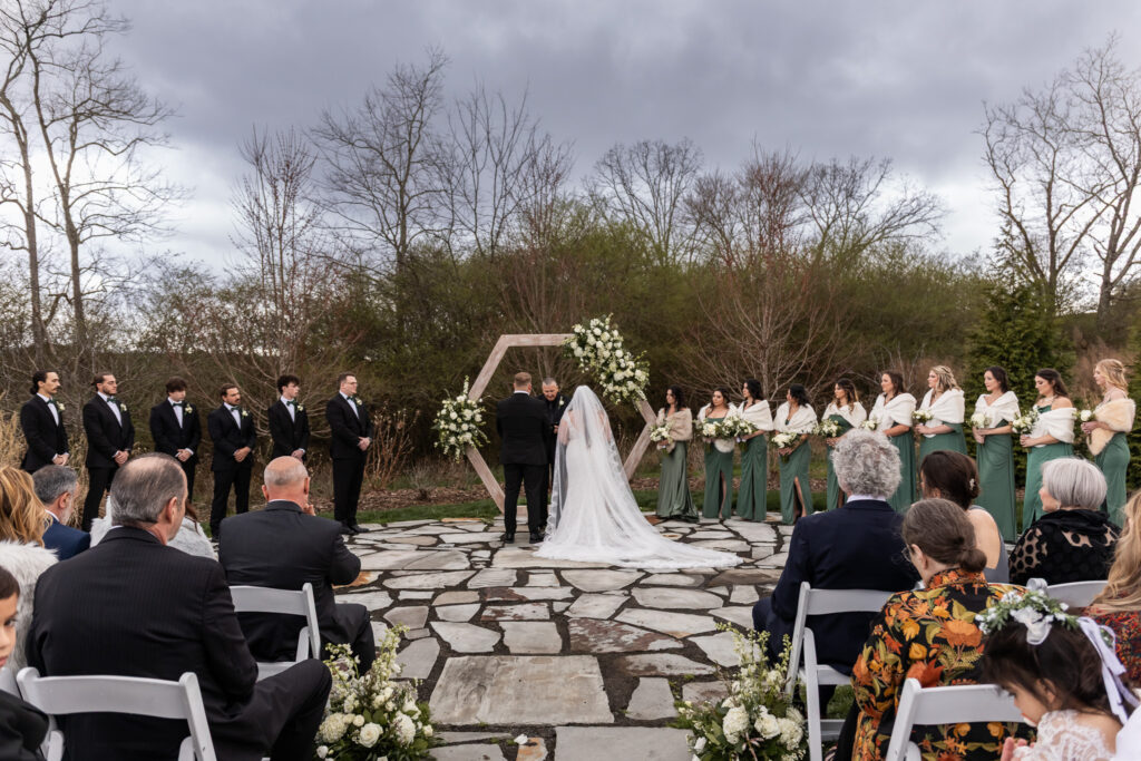 An outdoor ceremony shows the couple standing at the center before a wooden hexagon arch adorned with white florals, their wedding party lined on either side in green gowns and black suits, all under a dramatic, cloud-filled sky that hints at the glow of sunset behind the trees.