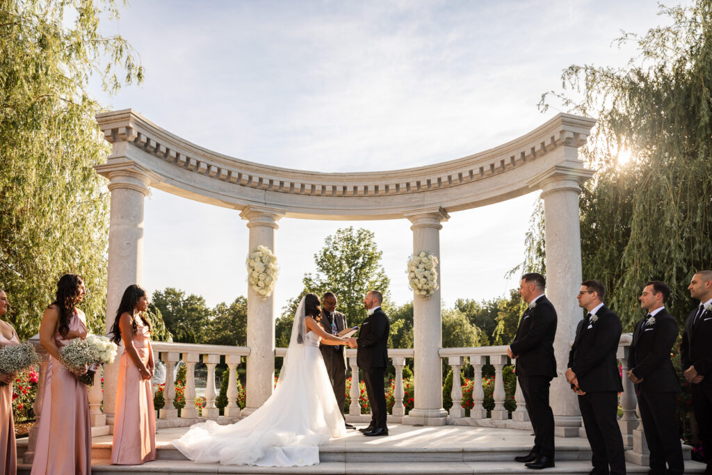A bride and groom stand beneath a curved colonnade of white stone pillars with their wedding party arranged on either side, soft afternoon sun glowing behind the structure and greenery, offering a clear example of what time do weddings usually start when planning elegant outdoor portraits with directional light.