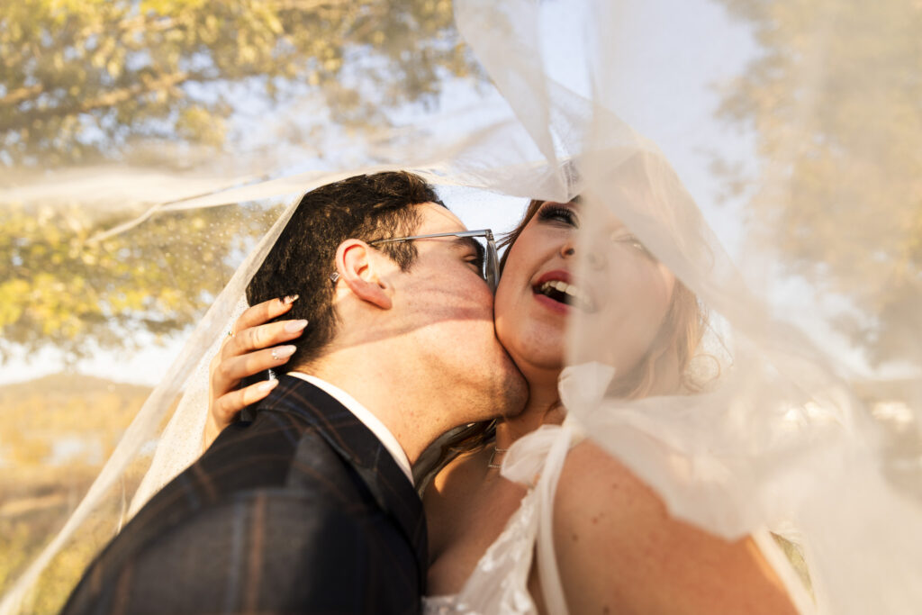 A close, intimate portrait of a couple beneath the bride’s veil shows the groom kissing her neck while warm golden light glows through the fabric around them, with soft autumn trees blurred in the background and the word sunset fitting the mood of this romantic, glowing moment.