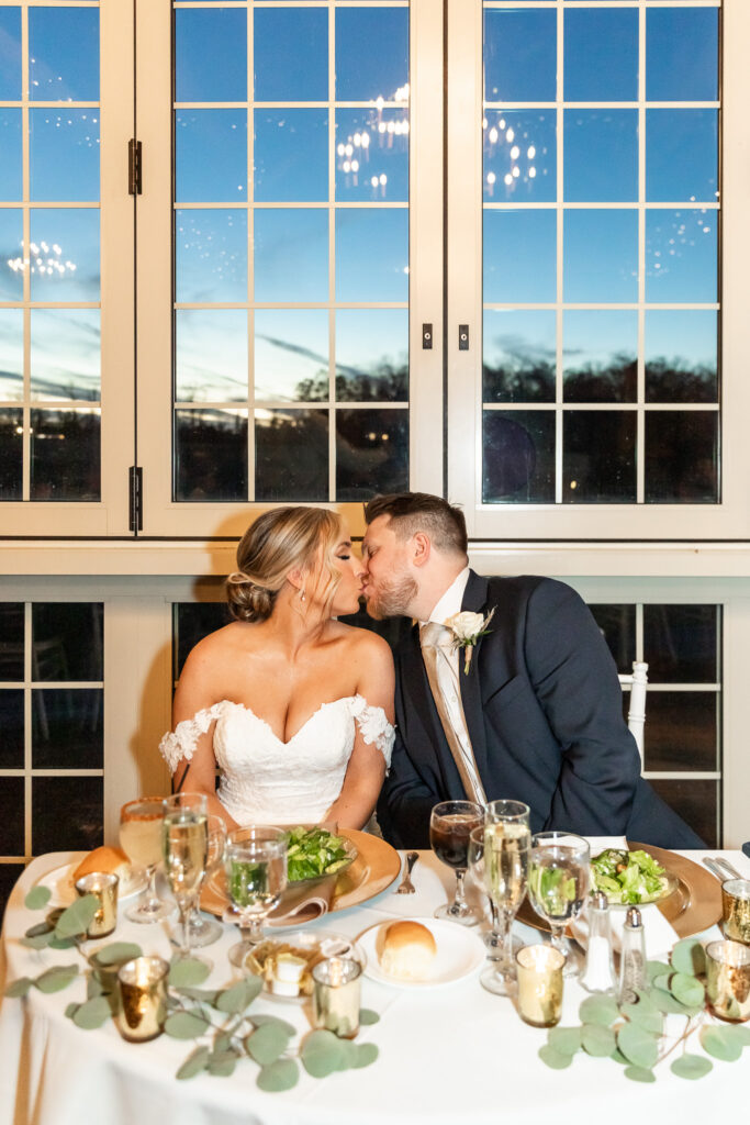 A couple shares a kiss at their sweetheart table inside a reception venue, their plates, champagne flutes, and greenery spread across the table while large windows behind them reveal the deepening blue sky just after sunset.