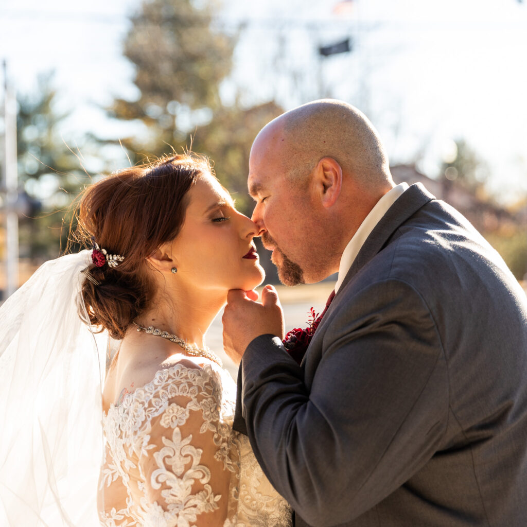 A bride and groom lean in for a soft forehead-to-nose kiss, her lace sleeves and veil glowing in bright backlight while the groom gently lifts her chin, all set against a warm afternoon that transitions toward sunset in the background.