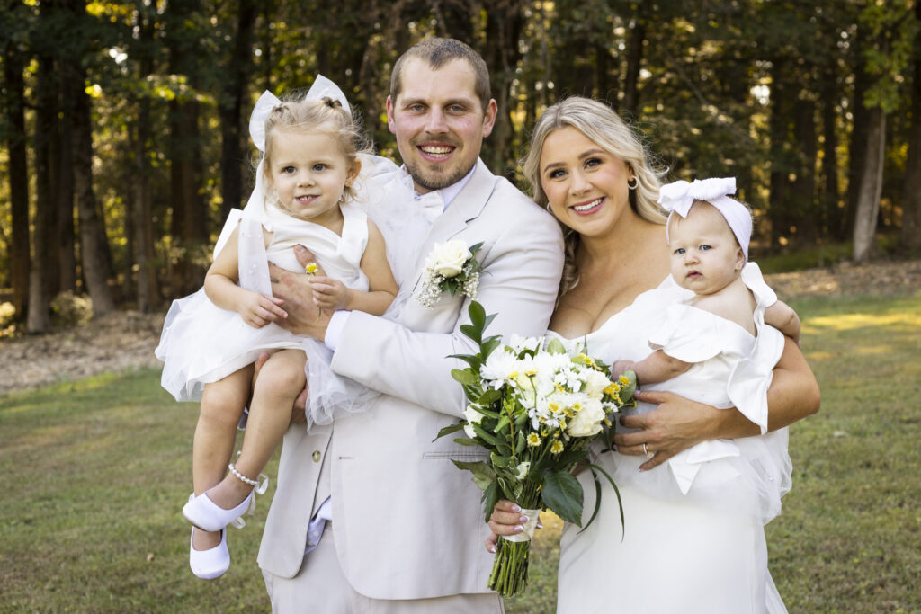 Bride and Groom hold their two young daughers in front of tree line with golden light peeking through
