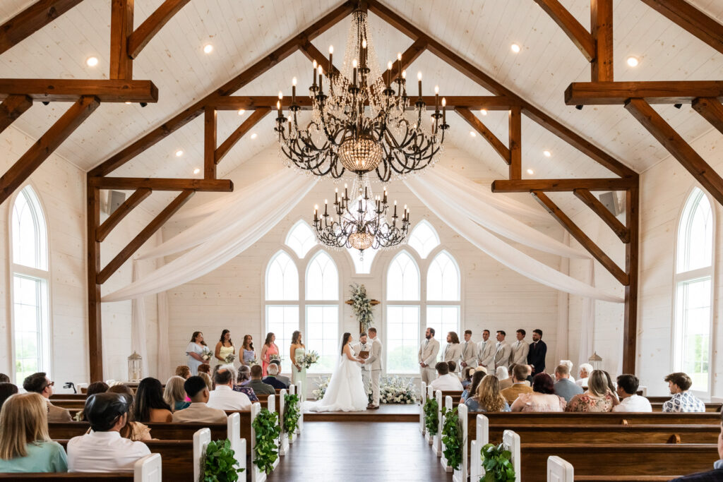 Inside a white-walled chapel with soaring wood beams and chandeliers, a couple exchanges vows at the front altar while guests fill wooden pews, the even indoor lighting providing a timeless backdrop that complements guidance on what time do weddings usually start for chapel ceremonies.