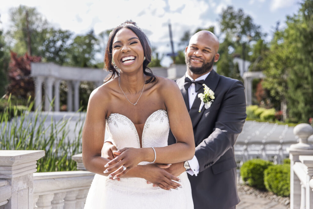 A joyful bride laughs as her groom wraps his arms around her from behind, both standing on a stone terrace with white pillars and lush greenery around them, the bright sky above showing early evening light before sunset.