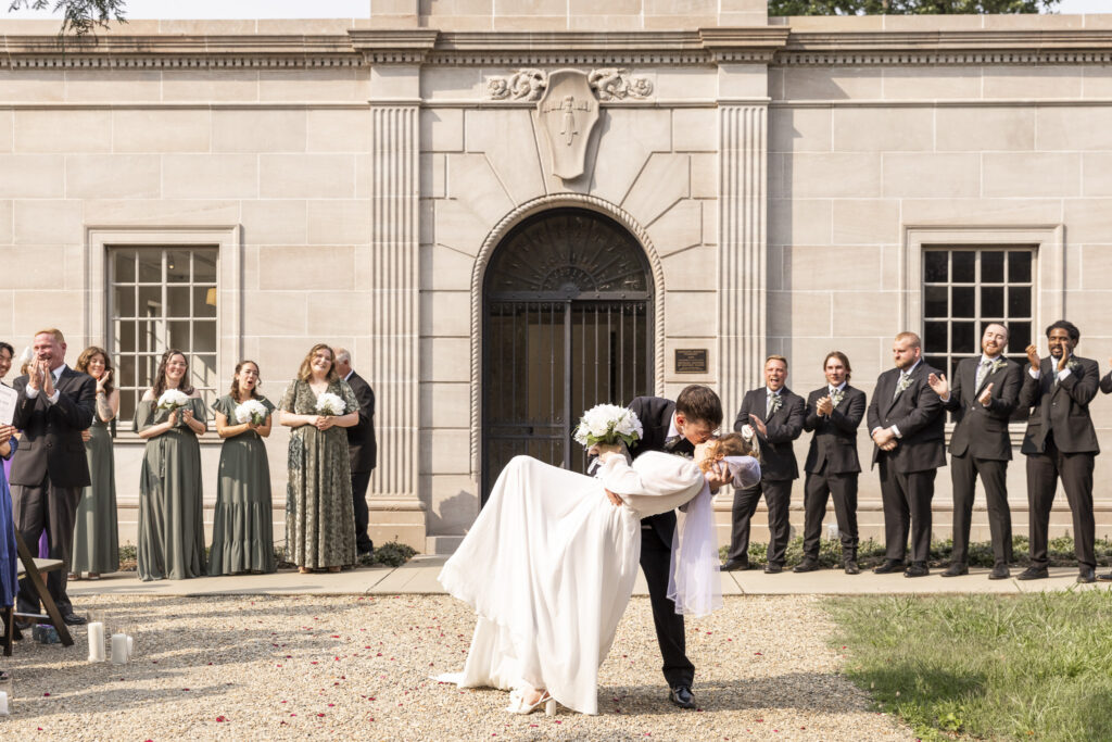 A groom dips his bride into a celebratory kiss in front of a stately stone building, surrounded by clapping wedding party members in sage dresses and black suits, the bright midday sun casting clean shadows that demonstrate what time do weddings usually start for a classic daytime ceremony.