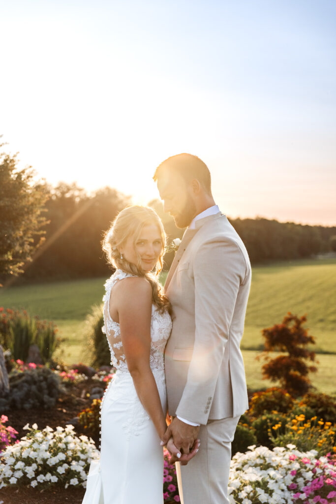 A bride and groom stand close together in a garden at sunset, golden light wrapping around them as the bride turns toward the camera and flowers bloom at their feet, creating a romantic portrait that highlights what time do weddings usually start when couples want those glowing end-of-day images.