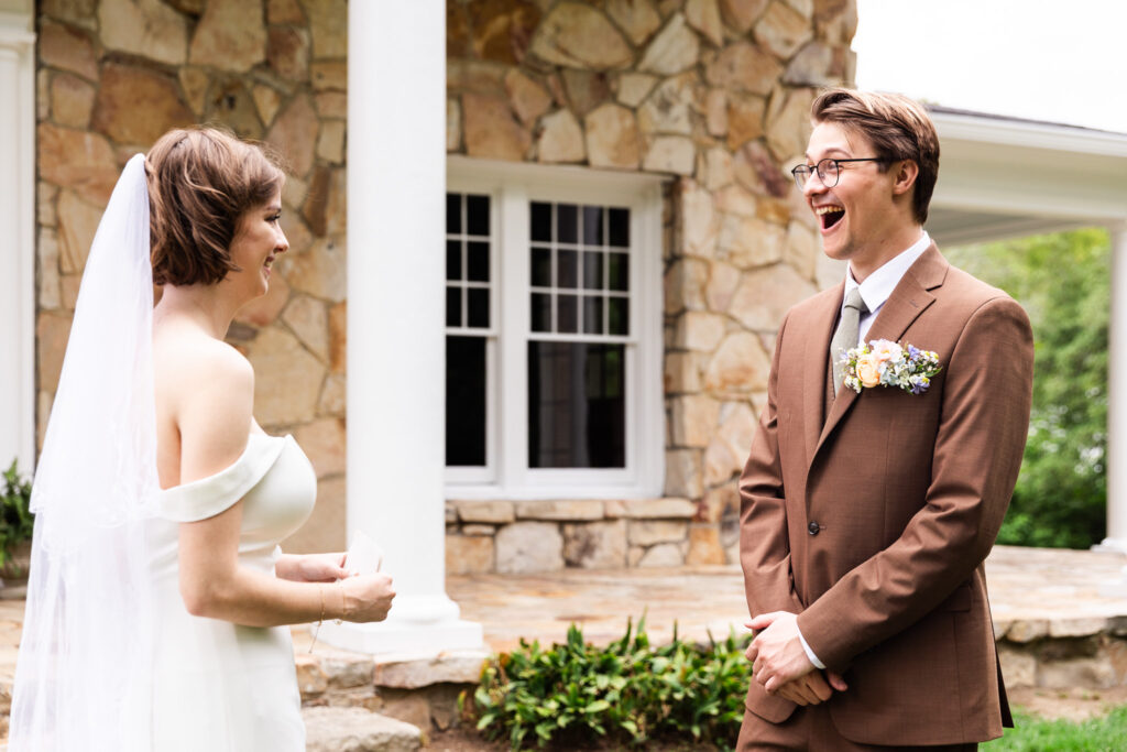 A bride and groom see each other for the first time outside a stone-front house, the bride smiling with her bouquet tucked at her side while the groom reacts with wide-open excitement, his hands clasped in front of him as he stands in a brown suit beneath white porch columns.