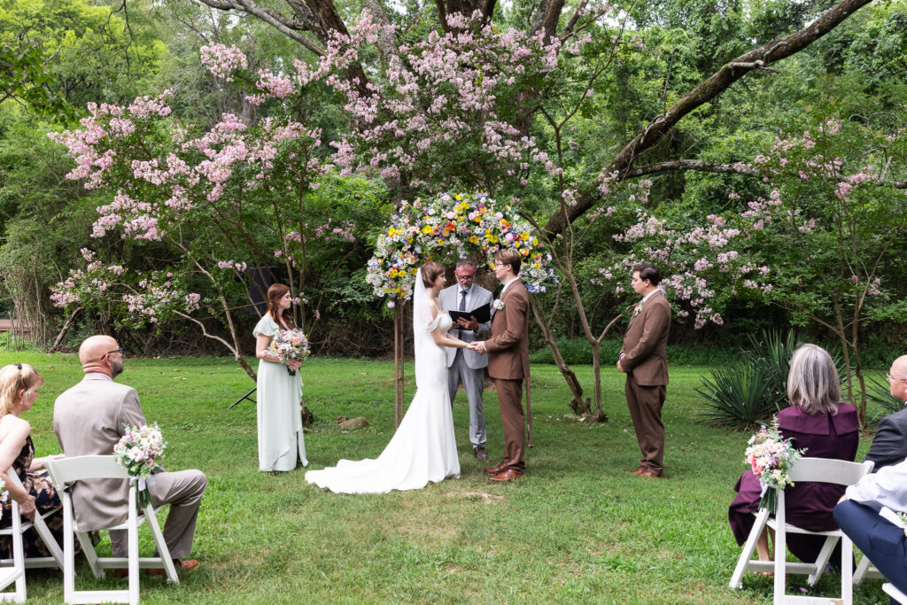 An outdoor ceremony takes place beneath soft pink flowering trees, with the couple standing under a colorful floral arch as guests watch from white chairs, the lush greenery and evenly lit moment showing what time do weddings usually start when choosing a springtime celebration with gentle daylight.