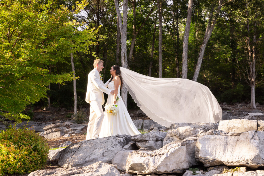 A bride and groom stand on sunlit rocks beneath tall green trees, the bride’s veil sweeping dramatically behind her as she holds a yellow-and-white bouquet and smiles toward her partner, all captured in warm golden light that highlights what time do weddings usually start for couples hoping for bright afternoon portraits.