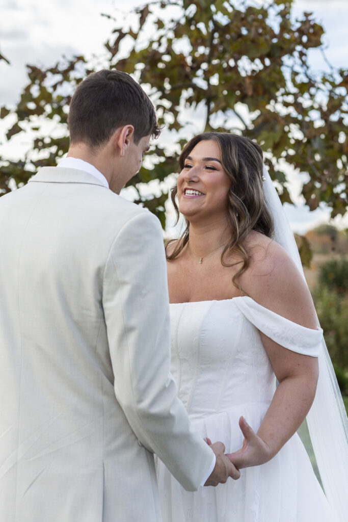 The bride holds her groom’s hands and beams up at him, her face glowing with emotion as early autumn leaves frame the sky behind them in the middle of a garden party wedding, creating a moment that feels warm, grounded, and completely theirs.