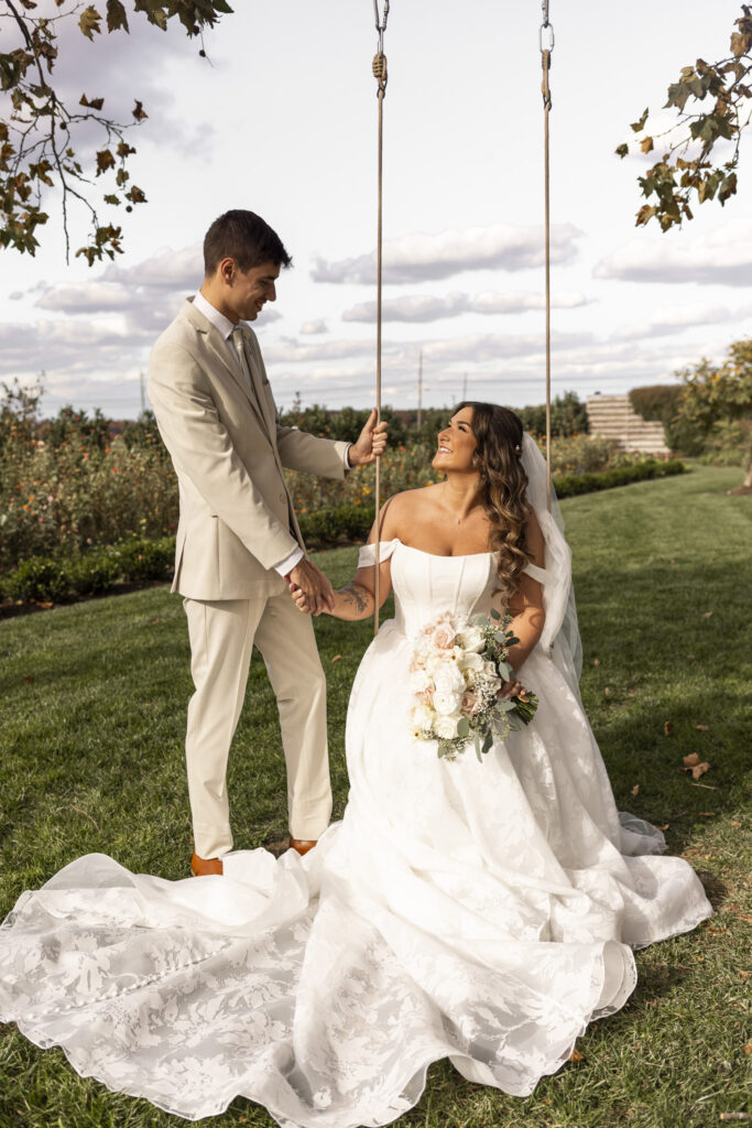 Outside on a lush lawn, the groom steadies a wooden swing while the bride sits gracefully in her voluminous gown, looking up at him with a bright, joyful smile. Her bouquet of cream and blush flowers rests in her lap, catching the afternoon sunlight, and behind them stretches a landscape of trimmed gardens and open fields that echo the pastoral beauty found across south jersey rustic wedding venues known for their wide outdoor spaces. The scene feels playful and romantic, framed by autumn leaves overhead.