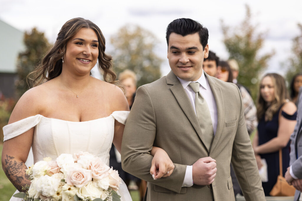 The bride walks down the aisle linked arm-in-arm with her escort, carrying a bouquet of soft blush and ivory blooms as guests rise around her, the outdoor setting capturing the gentle romance of a garden party wedding just before the ceremony begins.
