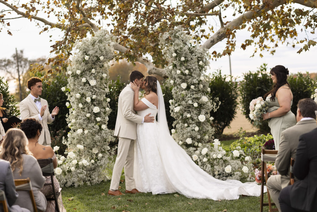 The couple shares their first kiss between two grand floral pillars made of baby’s breath and white roses, set beneath a canopy of autumn branches. Guests seated outdoors watch the moment unfold, and warm late-day sunlight softens the scene. The ceremony’s aesthetic—romantic florals, open air, and understated elegance—aligns beautifully with the style of south jersey rustic wedding venues, known for blending pastoral scenery with refined design elements.