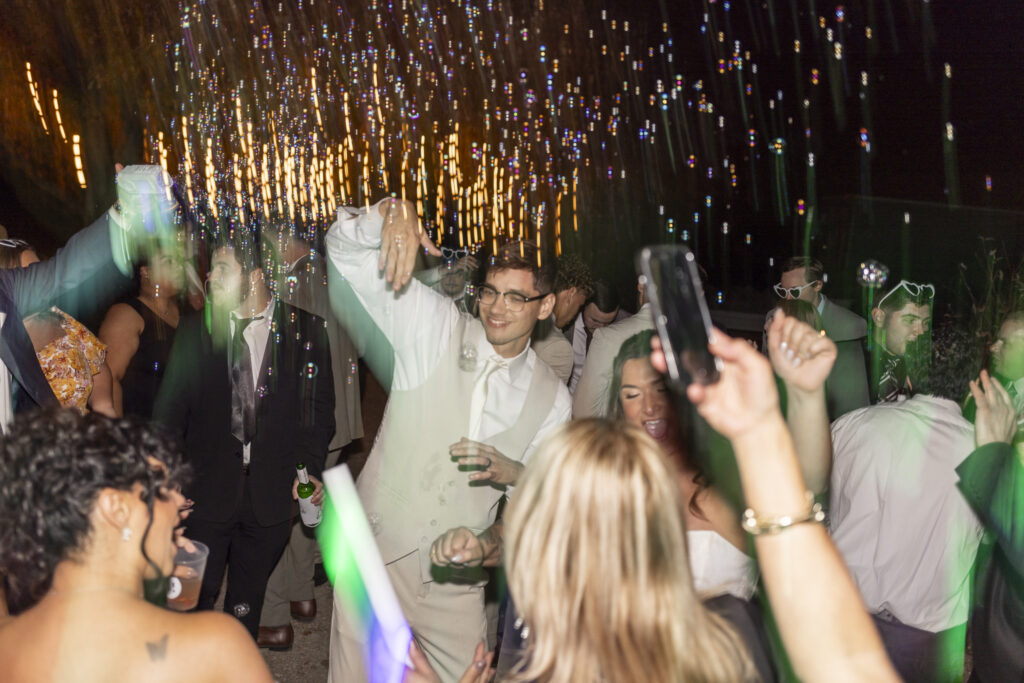 The groom dances in the middle of a packed outdoor dance floor, his arm raised as bubbles drift through warm string lights overhead. Guests surround him with raised hands, drinks, and glow sticks, their faces lit with excitement. The motion blur from the lights adds vibrant streaks of color, capturing the wild energy of the reception.