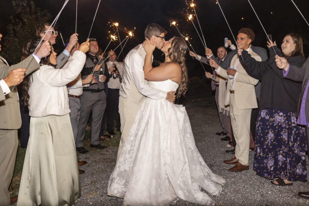 The couple shares a kiss beneath a glowing archway of sparklers, her dress sweeping wide as guests cheer from both sides, creating a magical nighttime finale to a garden party wedding filled with warmth and celebration.