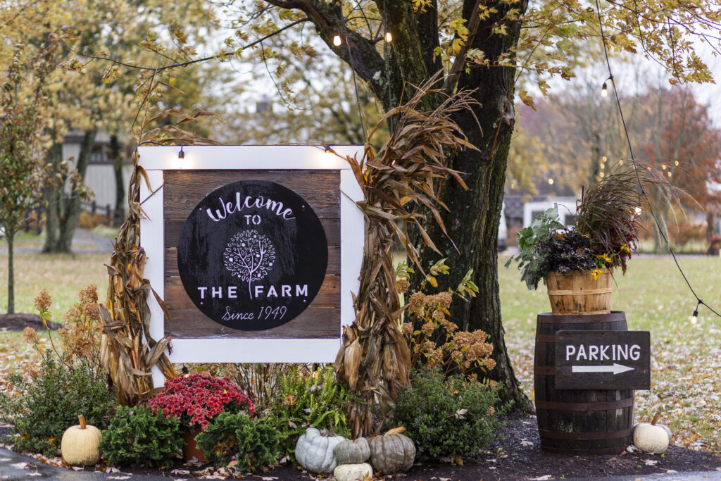 A wooden “Welcome to The Farm” sign is decorated with cornstalks, mums, pumpkins, and string lights, and the cozy, harvest-inspired display sits beneath a tree whose branches sway in the breeze—an inviting, seasonal entrance that suits the relaxed charm of a bucks county wedding.