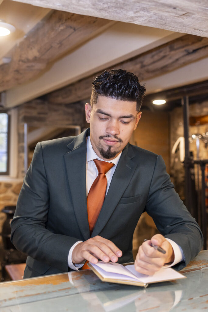 The groom leans over a rustic wooden counter inside a stone-walled room, writing carefully in a small notebook while dressed in a deep green suit and orange tie, the warm overhead lights adding an intimate calm that feels fitting for a thoughtful moment during a bucks county wedding morning.