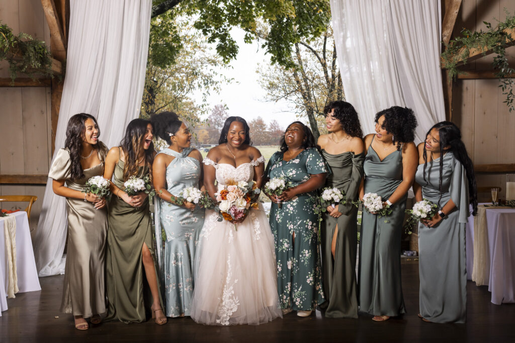 The bride stands surrounded by her bridesmaids inside a barn draped with white fabric, everyone laughing and leaning toward each other while holding small white bouquets, and the combination of muted green dresses and fall foliage outside creates a lively, celebratory moment that embodies the warmth of a bucks county wedding.