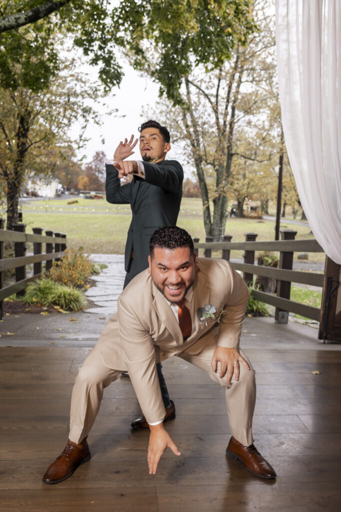Two groomsmen strike exaggerated, comedic poses on the wooden pavilion floor while the fall landscape stretches out behind them, and their laughter-filled antics add a lighthearted pause in the portrait session typical of a joyful The Farm Bakery and Events wedding.