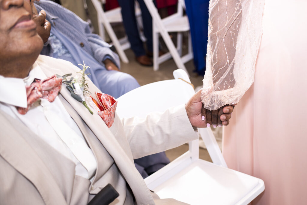 A close-up of two hands joined tightly during the ceremony shows an older man in a light beige suit holding the hand of a woman in a pale pink gown (the brides parents). His bow tie and pocket square are a vibrant coral pattern, and their fingers are interlaced tenderly as they sit among guests in white folding chairs.