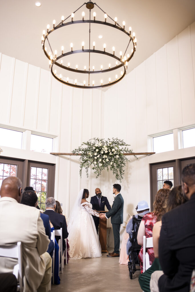 The couple stands hand-in-hand at the front of an indoor ceremony space with tall white walls and windows. The officiant stands between them beneath a hanging arrangement of white flowers and greenery, while guests watch from rows of white chairs below a large circular chandelier glowing overhead.