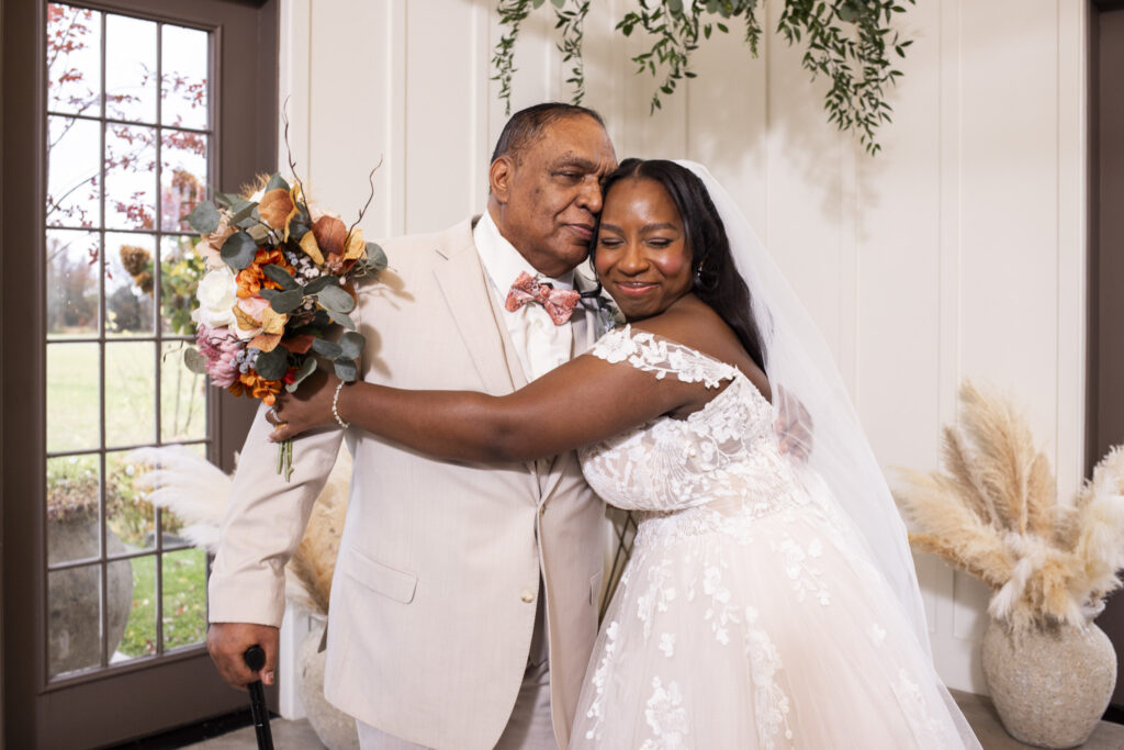 The bride wraps her arms around her dad dressed in a beige suit, leaning her head into his cheek with a peaceful smile. He holds a cane in one hand while the other rests around her back, and they stand together in front of tall windows with soft pampas arrangements behind them.
