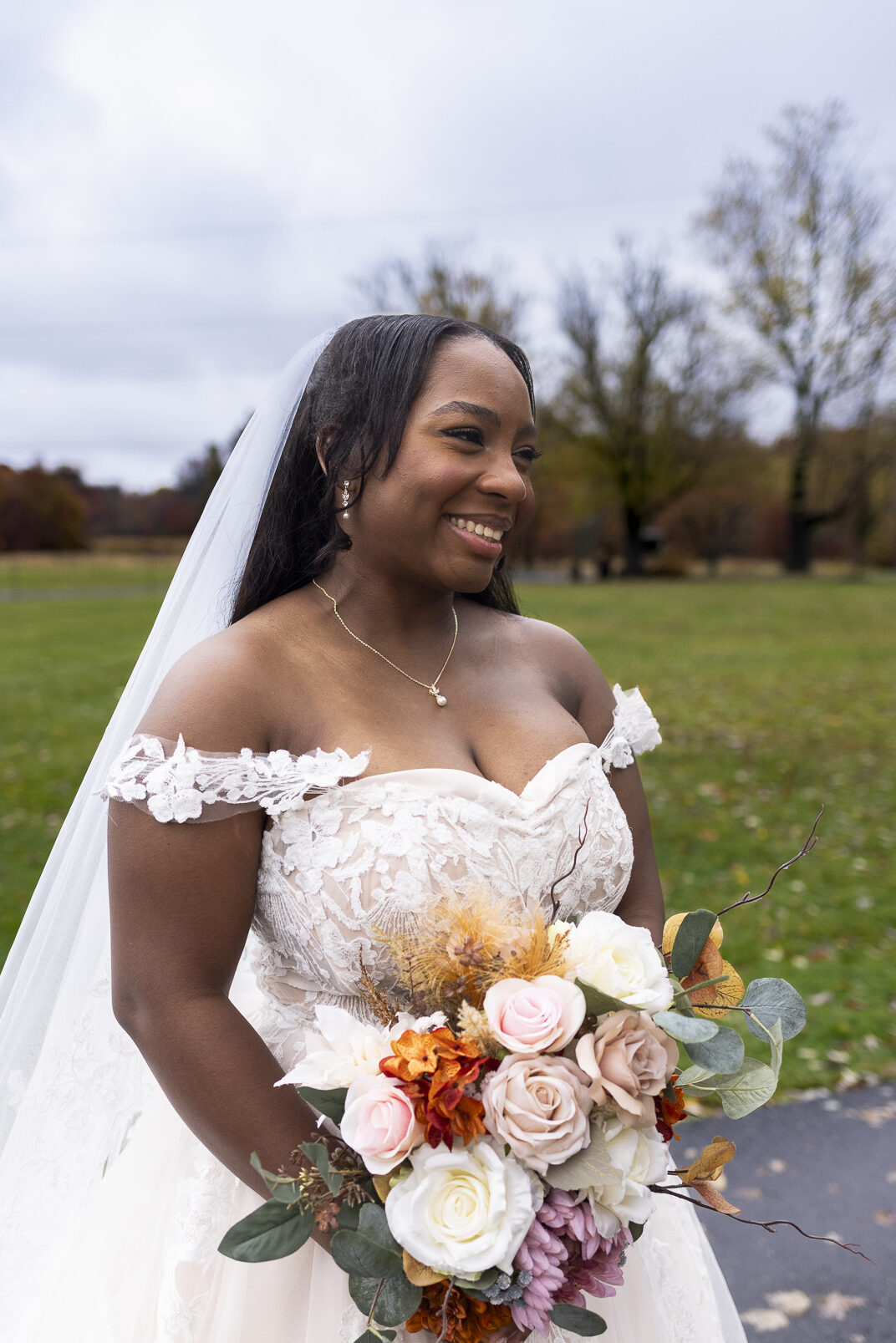 The bride stands outdoors holding her bouquet of soft pink, cream, and copper-toned flowers, smiling toward the distance as the muted fall landscape behind her reflects the calm beauty of The Farm Bakery and Events wedding on an overcast afternoon.