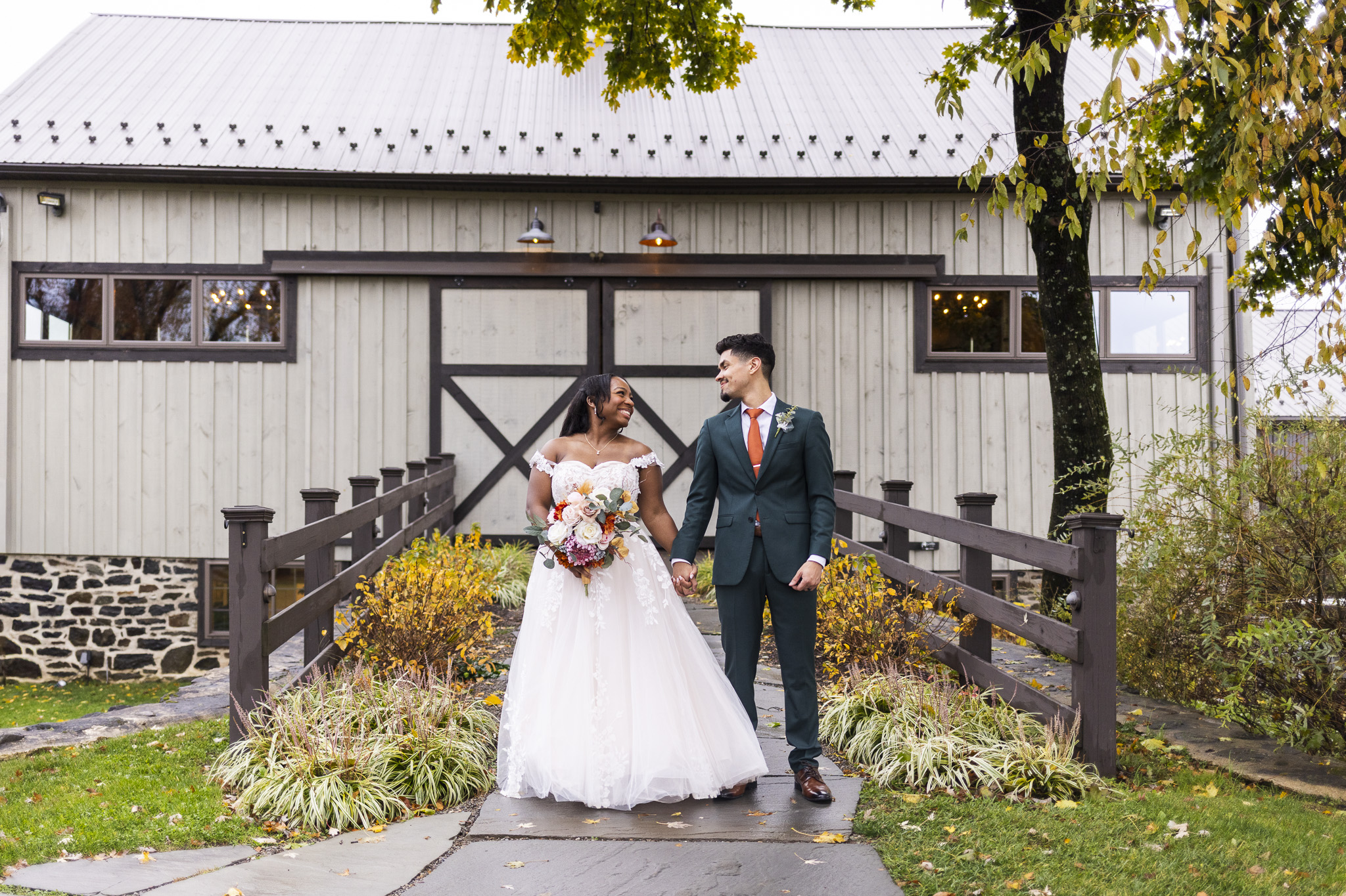 The couple stands hand-in-hand on a stone path leading up to a rustic barn, smiling warmly at each other as soft fall foliage surrounds them, and the scene captures the relaxed charm of a bucks county wedding with golden leaves, wood railings, and muted gray barn siding framing the moment.